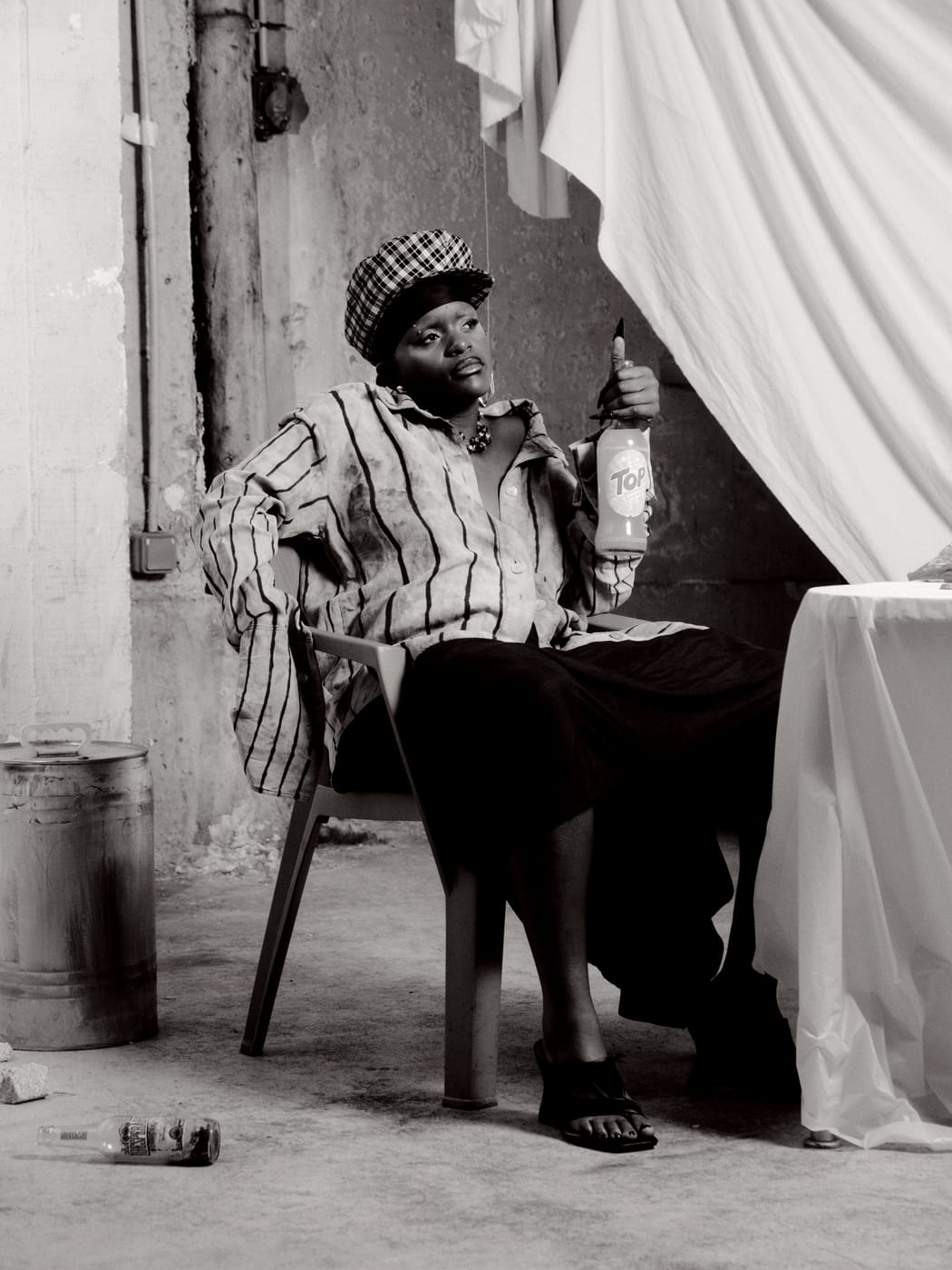 Ezek seated at the table in the beret, a bottle in one hand, toned monochrome