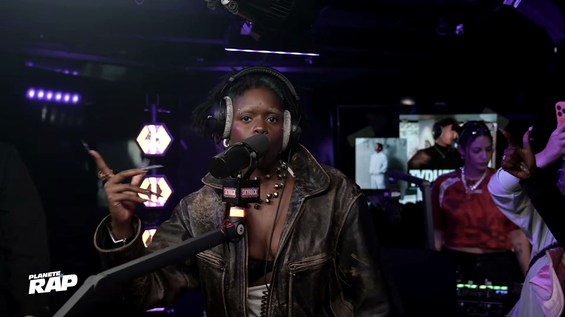 Planète Rap still: Ezek at the mic in a worn leather jacket, one hand raised, the studio in purple light