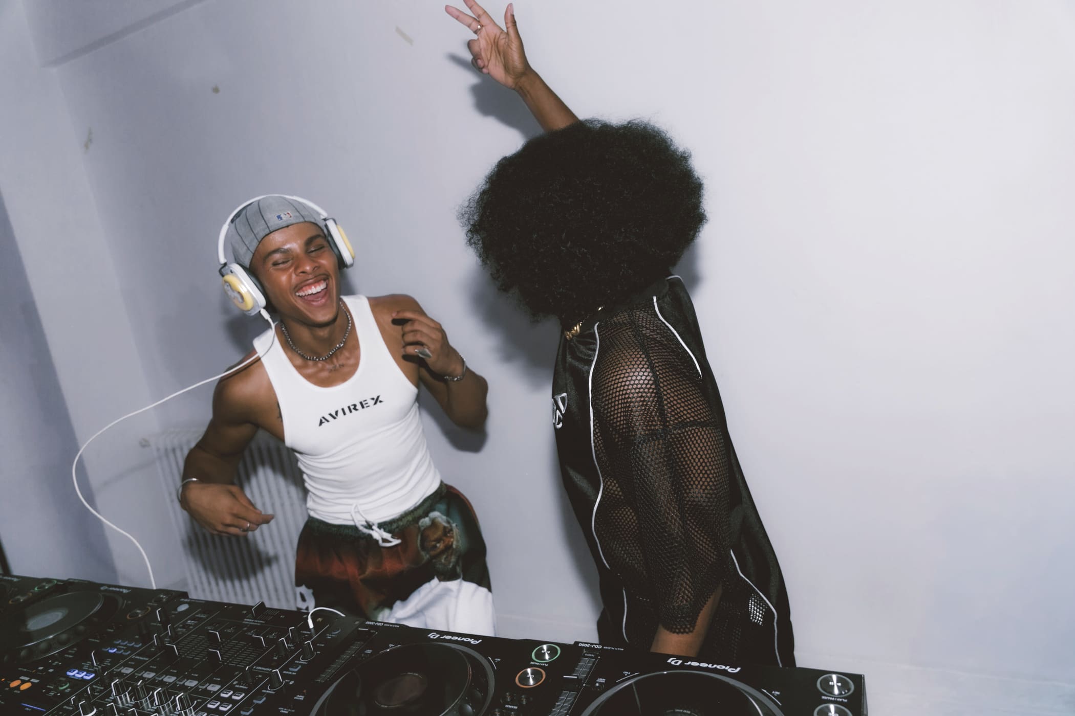 Oluwa laughing at the decks in cap and headphones, a dancer's arm thrown up beside him