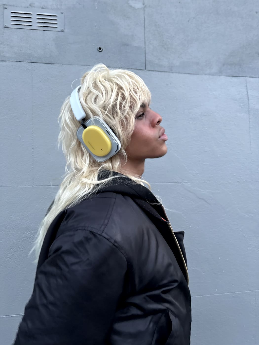 Oluwa in profile wearing Nothing headphones, blonde mullet against a grey wall